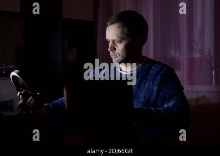 A man in home clothes works at a laptop. Sits in the twilight, puts headphones on his head. Stock Photo