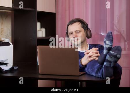 A man in home clothes works at a laptop. Headphones are on the head. He put his feet on the table. Stock Photo