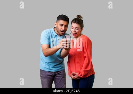 Friends reading shocking news! Portrait of amazed attractive young couple in casual wear reading unexpected message with open mouth, using mobile phon Stock Photo
