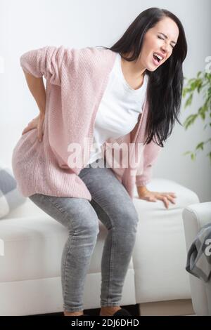 Intense pain in lower back holds a woman from straightening when trying to stand up, having painful expression on her face. Stock Photo