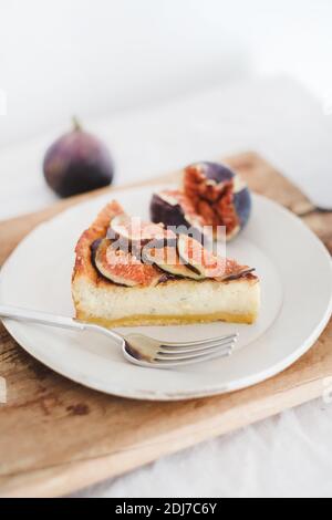 Slice of homemade gluten free fig cheesecake with fresh seasonal figs pieces on white plate over rustic wooden board, white wall at background. Healthy dessert, artisan sweet food concept Stock Photo