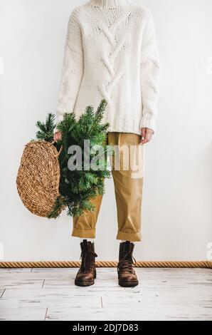 Human figure in white woolen sweater, khaki pants and boots standing holding wicker bag with evergreen Christmas tree branches for winter holidays celebration at home. Christmas, New year concept Stock Photo