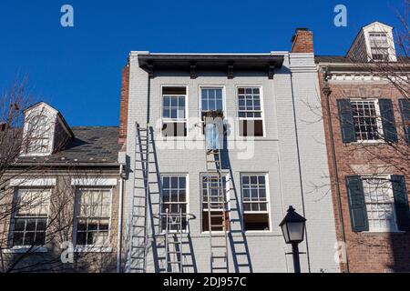 A painter is on top of an industrial size aluminum extension ladder painting the exterior surface of an old brick house. He wears no safety equipment Stock Photo