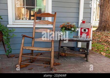 A porch decoration at a colonial era house featuring an old wooden rocking chair, a small table with metal blacksmith tools and a model farmhouse on t Stock Photo