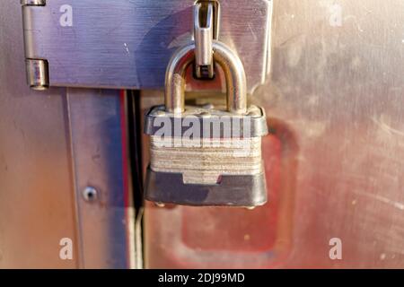 Close up isolated image of a cast iron rough textured heavy duty padlock attached securely on a hinge door of a metal cabinet. Sunlight reflects from Stock Photo
