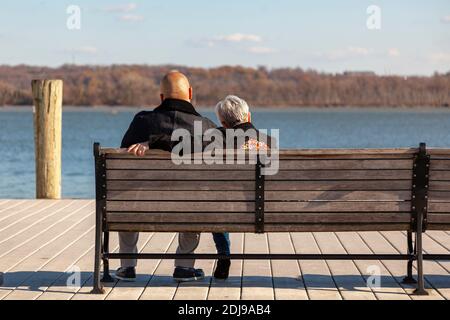 an elderly caucasian woman with short gray hair and a younger African American man with shaved head are sitting on a bench on a pier looking at sea. I Stock Photo