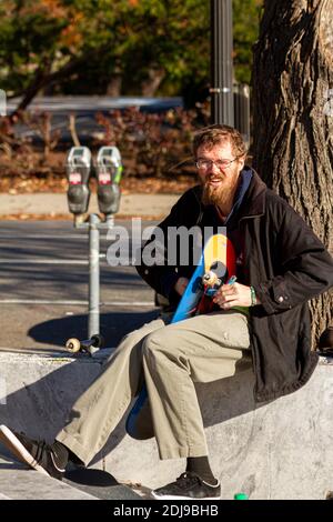 Washington DC, USA 11-28-2020: A young caucasian man with red goatee beard is sitting on a wall assembling his skateboard. He is screwing in the truck Stock Photo