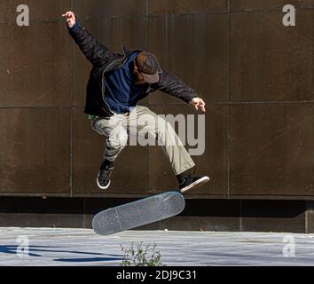 A young bearded caucasian man wearing baseball hat, canvas pants and a thin coat is doing a 360 rotation with his skateboard. He jumps in the air with Stock Photo