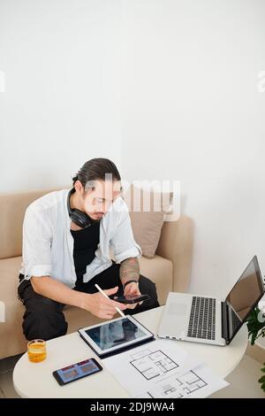 Serious interior designer checking text messages from client when working with plans and images of his house Stock Photo