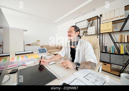 Smiling mixed-race designer using digital pen when drawing sketch for client at his desk in home office Stock Photo