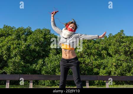 A Caucasian female with headphones joyfully jumping in a park Stock Photo