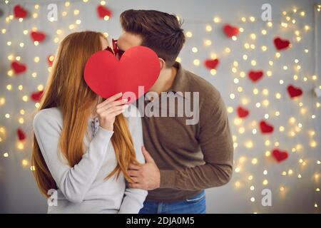 Unknown young man and woman kissing hiding behind a big red paper heart they are holding in hand. Stock Photo
