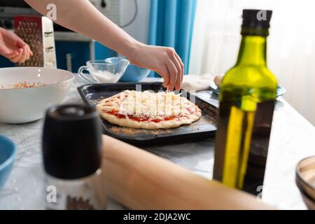 A female's hand sprinkles cheese on a pizza tray. Close-up. Side view. Home cooking. Stock Photo