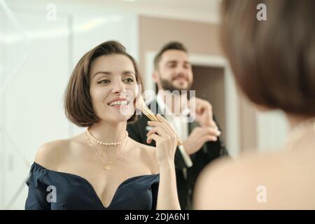 Happy gorgeous young woman applying makeup with brush in front of mirror while her boyfriend adjusting shirt sleeve Stock Photo