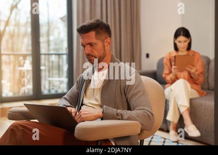 Portrait of modern couple working from home in minimal interior, focus on handsome mature man using laptop while sitting in ergonomic armchair Stock Photo