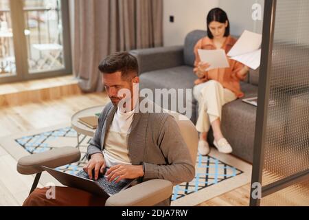High angle portrait of modern couple working from home in minimal interior, focus on handsome mature man using laptop while sitting in ergonomic armch Stock Photo