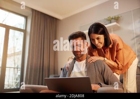 Portrait of handsome mature man using laptop while working from home with smiling wife embracing him, copy space Stock Photo