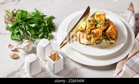 Strata or italian breakfast casserole of spinach, cheese and soaked overnight cubed bread baked with mustard on a white plate with golden cutlery on a Stock Photo