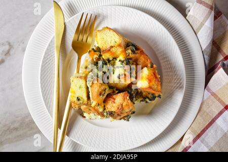 Strata or italian breakfast casserole of spinach, cheese and soaked overnight cubed bread baked with mustard on a white plate with golden cutlery on a Stock Photo