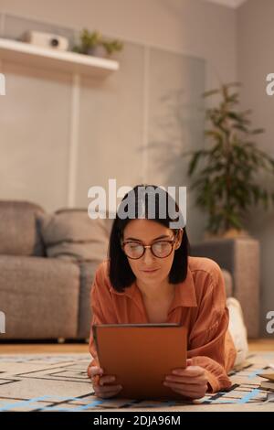Vertical portrait of contemporary woman working from home while lying on floor and using digital tablet, copy space Stock Photo