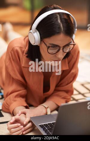 High angle portrait of contemporary woman working from home while lying on floor wearing headphones and using laptop Stock Photo