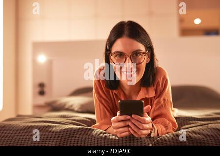 Portrait of contemporary adult woman using smartphone while lying on bed in minimal interior and smiling at camera, copy space Stock Photo