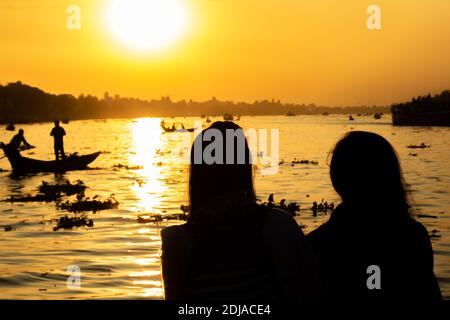 Silhouette of two girls looking at the Buriganga river at the sunset, with tens boats crossing the river in Dhaka. Stock Photo