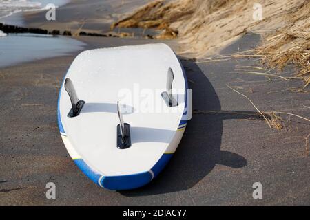 Wet bright sup-board lying on the sandy beach with black fins up. Closeup. Drops of water on the surf the desk. Surf in the background. USA, Michigan, Stock Photo