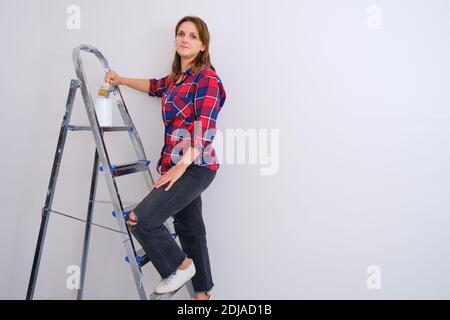 Woman painter stands on a stepladder in a plaid shirt, copy space. Painting the walls in a new apartment Stock Photo