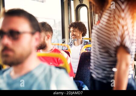Young cheerful handsome man is sitting on a bus looking through a window and smiling while being surrounded by other passengers. Stock Photo