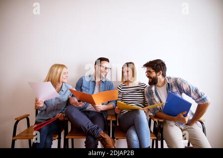 Group of young joyful satisfied friends holding colourful folder while sitting in a chair sharing notes and laughing in the waiting room for an exam. Stock Photo