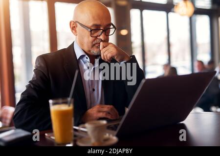 Mature professional worried businessman is brainstorming how to solve a problem while using laptop drinking coffee and juice in coffee shop. Stock Photo
