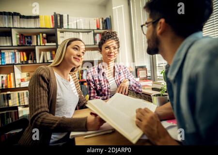 Two young focused cheerful female students are listening to the lecturer and taking notes while sitting at the desk near window and bookshelf in the s Stock Photo