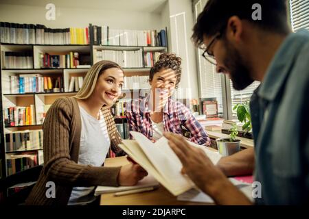 Two young joyful female students are smiling at and taking notes from a male lecturer while sitting at the desk near window and bookshelf in the schoo Stock Photo
