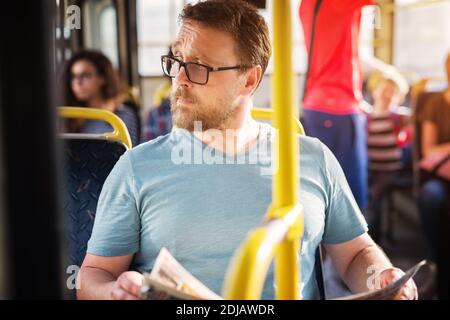 Young man is holding newspapers while looking out the bus window with his eyebrow raised. Stock Photo