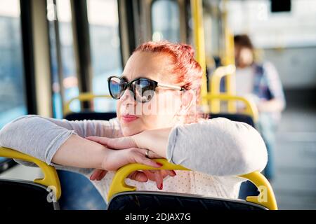 Mature woman with sunglasses on is sitting in a bus seat and leaning onto the seats in front of her and looking through the window. Stock Photo