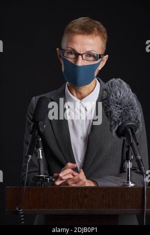 Vertical portrait of mature woman wearing mask while giving speech standing at podium against black background Stock Photo