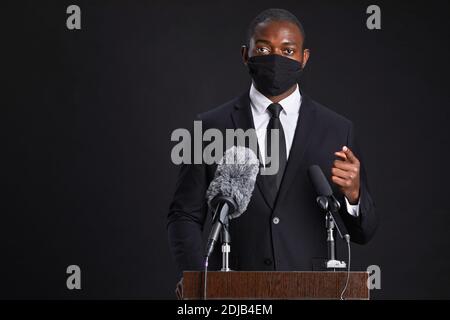 Waist up portrait of African-American man wearing mask while giving speech standing at podium against black background, copy space Stock Photo