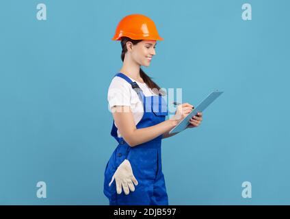 Smiling young handywoman in coveralls and hardhat making list of construction materials Stock Photo