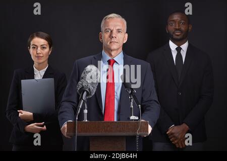 Waist up portrait of mature man giving speech standing at podium with two assistants in background Stock Photo