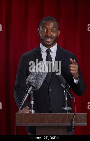 Vertical portrait of African-American man giving speech standing at podium on stage against red curtain Stock Photo