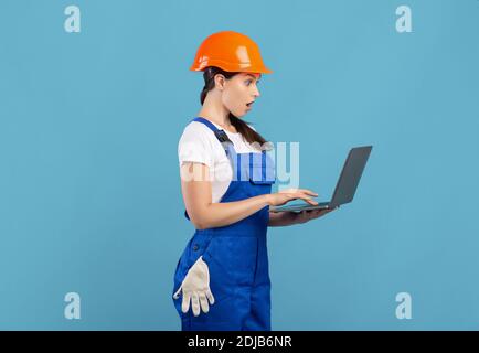 Shocked Young Handywoman In Coveralls And Hard Hat Using Laptop Computer Stock Photo