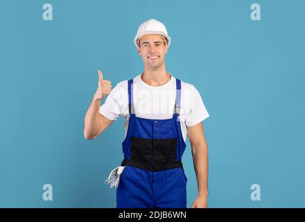 Portrait Of Young Maintenance Worker In Hardhat And Coveralls Showing Thumb Up Stock Photo