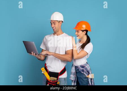 Young couple in hardhats using laptop during house repair, blue background Stock Photo