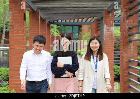 smiling Asian female and male college students with computer discuss and walk at a table in beautiful university campus with grass, tree and red brick Stock Photo