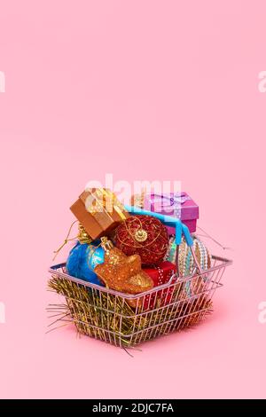 a metallic shopping basket full of different christmas ornaments and small gift boxes on a pink background, with some blank space on top Stock Photo
