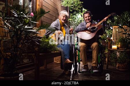 Senior couple in wheelchair with dog and guitar in the evening on terrace. Stock Photo