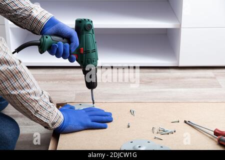 Man in gloves assembles table furniture with drill. Assembly, furniture repair home master. Male hand with drill on floor. Copy space. Stock Photo