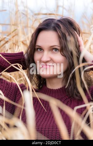 Portrait of beauty young woman in brown sweater posing in dried grass in field. Cute woman with long curly brunette hair and red lips. Stock Photo