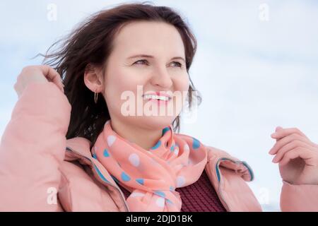 Happy young woman with beautiful smile and curly brunette hair fluttering in wind, dressed in beige jacket, scarf around her neck. Portrait of pretty Stock Photo